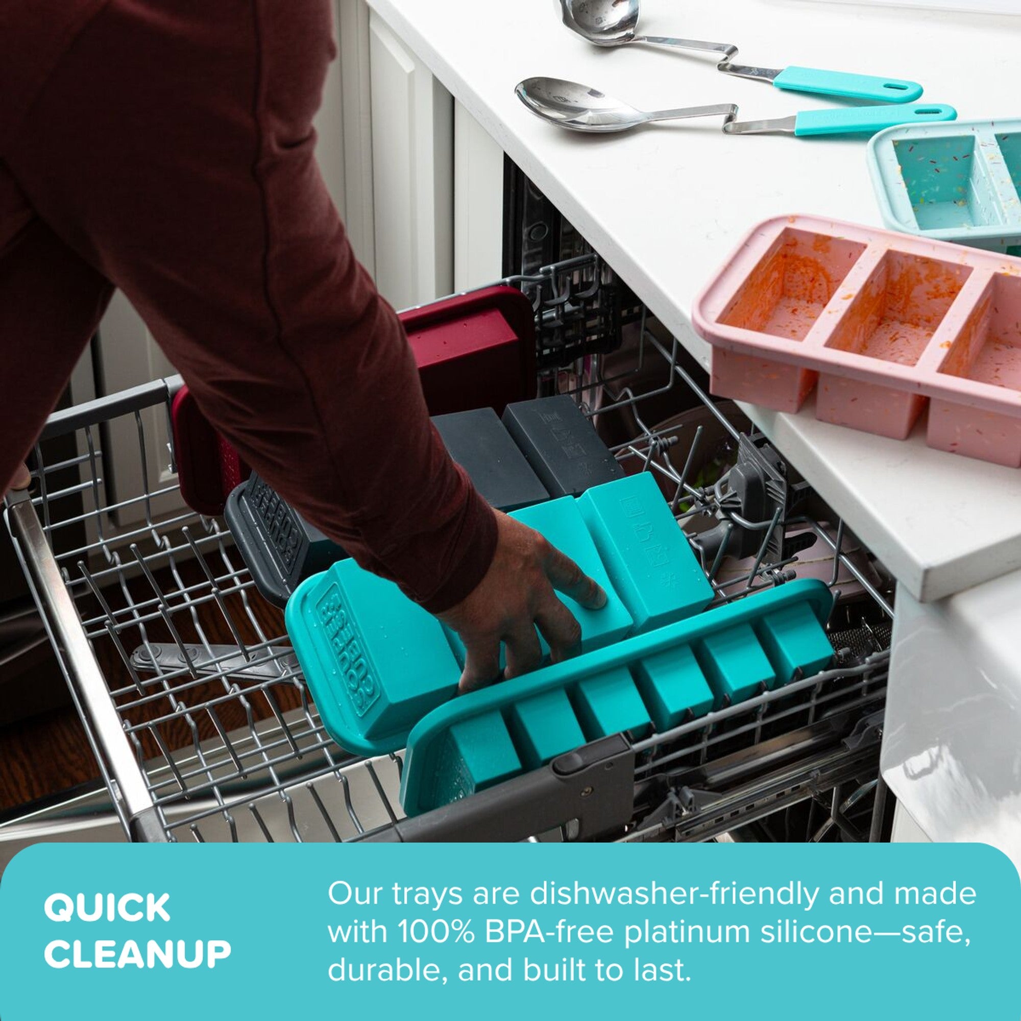 Person loading a teal silicone ice cube tray into a dishwasher with kitchen utensils and a pink ice cube tray on the counter.