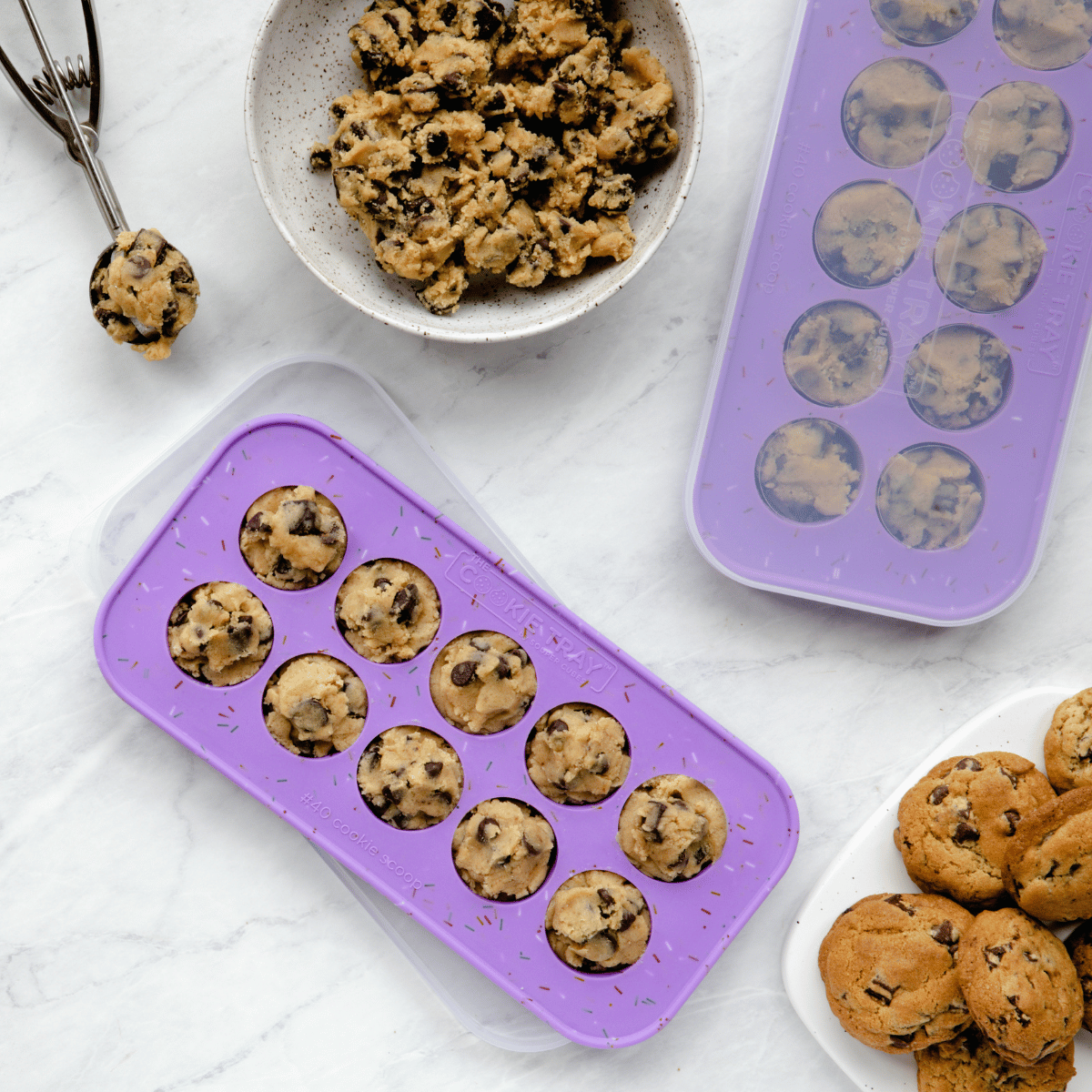 Purple silicone mold with cookie dough balls, a bowl of cookie dough, and a plate of baked cookies on a white surface.