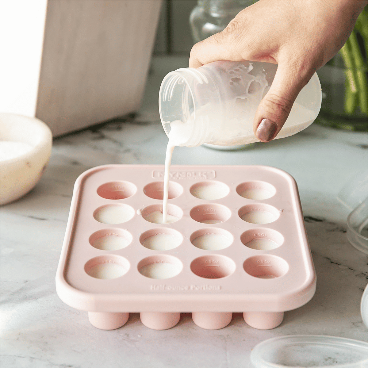 Pink silicone breast milk tray with a hand pouring milk into it on a marble countertop.