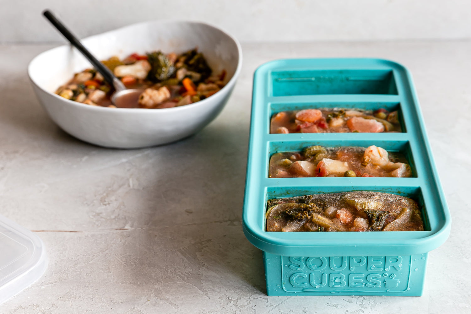½ cup Souper Cubes tray filled with frozen soup. Behind the tray is a white bowl filled with reheated soup.  