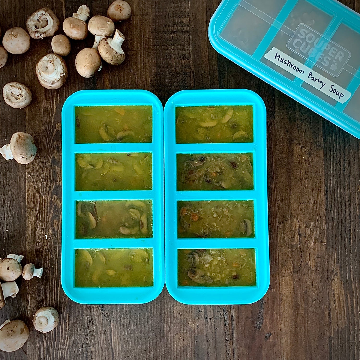 image of 3 1-cup souper cubes trays on a wooden table filled with mushroom barley soup. there are mushrooms along the left side of the picture.