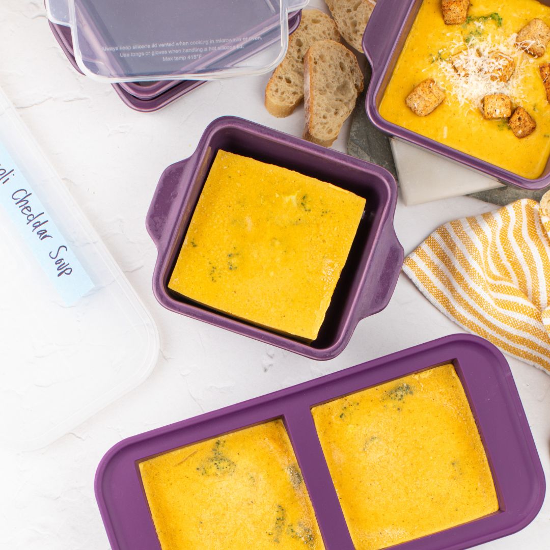 Purple silicone mold trays filled with baked cheddar soup, arranged on a white surface with bread, croutons, and stacked purple containers.