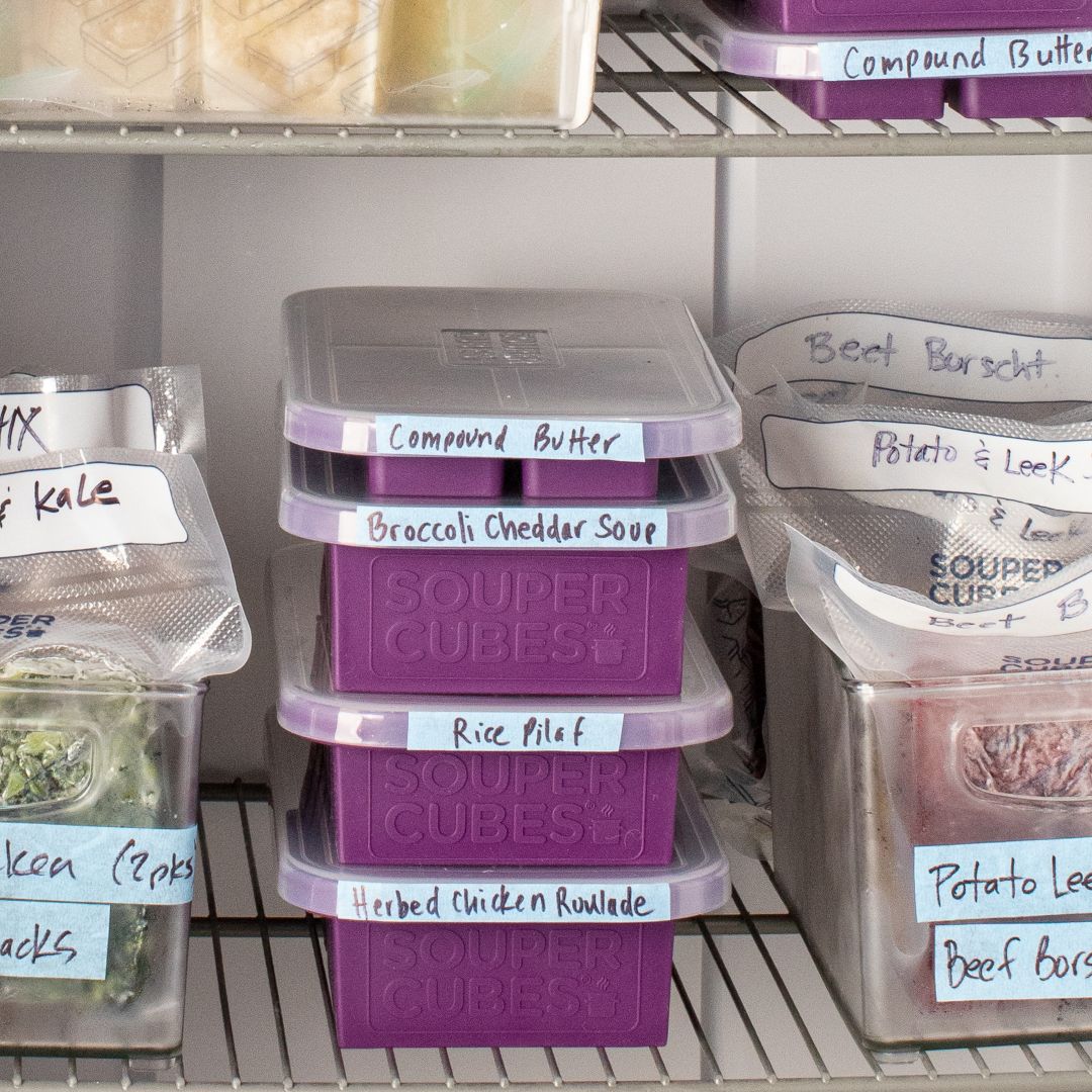A set of plum colored Souper Cubes freezer trays with clear lids, stacked on a  shelf alongside labeled containers of ingredients like compound butter, broccoli cheddar soup, rice pilaf, and potato leek.